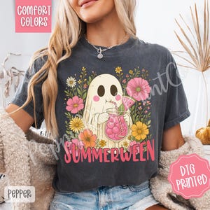 May include: Dark gray t-shirt featuring a cartoon ghost enjoying a pink drink with a watermelon slice, surrounded by flowers. The word "SUMMERWEEN" is printed in pink. The shirt has "COMFORT COLORS" and "DTG PRINTED" labels.