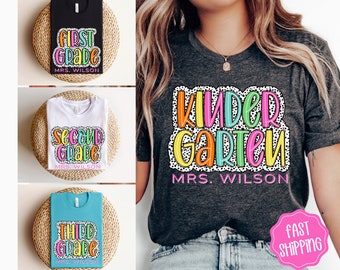 Może przedstawiać: T-shirty z wzorami dla poszczególnych klas. Koszulki są czarne, białe i turkusowe, każda z kolorową grafiką ze słowami "First Grade", "Second Grade" i "Third Grade", a także "MRS. WILSON". Szara koszulka wyświetla napis "Kindergarten MRS. WILSON".