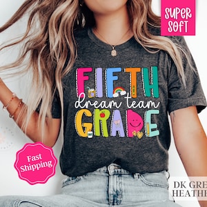 May include: A dark grey heather t-shirt with the text "FIFTH dream team GRADE" in colorful letters and illustrations. The text is surrounded by colorful illustrations of school supplies, a rainbow, and a smiley face.
