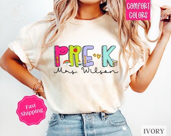 Peut inclure: T-shirt couleur ivoire avec le texte "PRE-K Mrs. Wilson" en lettres colorées avec des illustrations de fournitures scolaires et de cœurs.