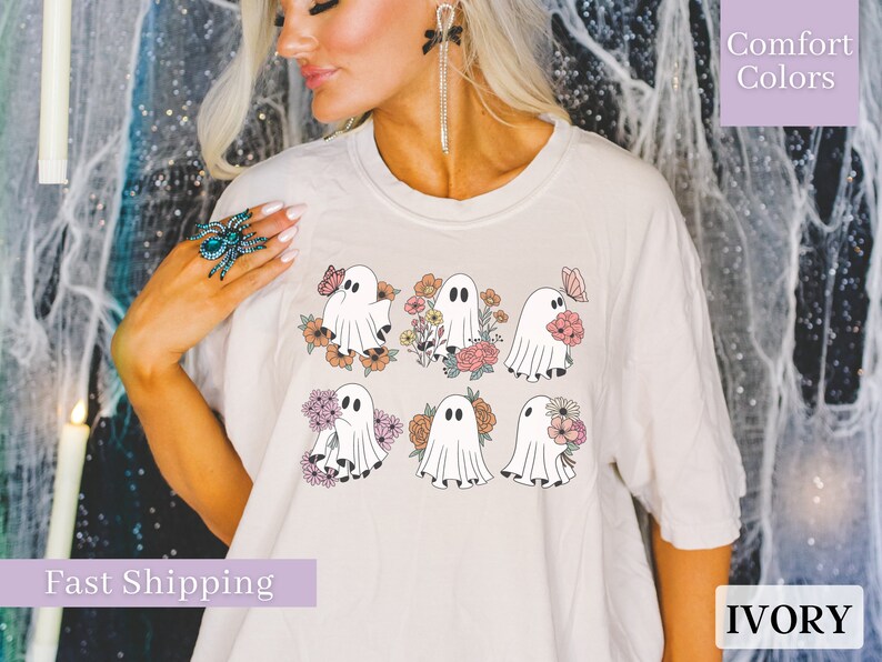 May include: A white t-shirt with a graphic of six ghosts holding flowers and butterflies. The ghosts are in a pastel color palette. The text "Comfort Colors" is in the upper right corner of the image. The text "IVORY" is in the lower right corner of the image.