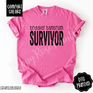 May include: A bright pink t-shirt with the words "toddler tantrum SURVIVOR" in black block letters. The shirt has short sleeves and a crew neck. The words "Comfort Colors" are in a black box in the upper left corner.