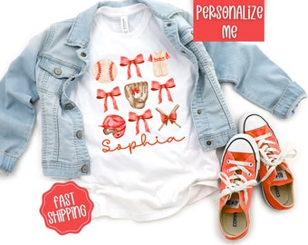 May include: White t-shirt with a baseball theme. The shirt features red bows, a baseball, a baseball glove, a baseball cap, and a baseball bat and ball. The name "Sophia" is written in cursive on the shirt.