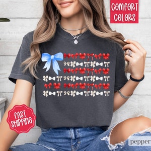 May include: Dark gray t-shirt featuring a watercolor design of the American flag made of bows. The flag design includes a blue bow in the upper left corner and rows of red, white, and blue bows. The shirt has the words "Comfort Colors" and "Fast Shipping" on it.