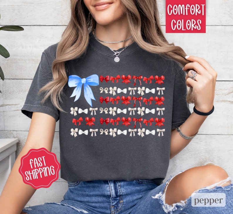 May include: Dark gray t-shirt featuring a watercolor design of the American flag made of bows. The flag design includes a blue bow in the upper left corner and rows of red, white, and blue bows. The shirt has the words "Comfort Colors" and "Fast Shipping" on it.