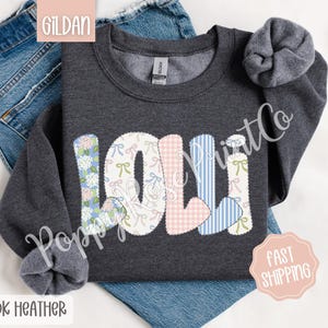 May include: Dark heather grey sweatshirt with the name "Lolli" spelled out in colorful patchwork letters. Each letter is decorated with a different floral pattern, including pink gingham, blue stripes, and white with pink bows.