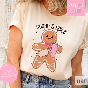 Op de afbeelding: Een beige t-shirt met de tekst "sugar & spice" en een cartoon peperkoekman die een roze beker vasthoudt. De peperkoekman draagt een roze strik.