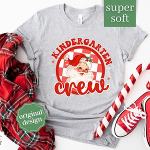May include: Gray t-shirt with a red and white checkered circle design featuring a cartoon Santa Claus and the text "Kindergarten Crew".