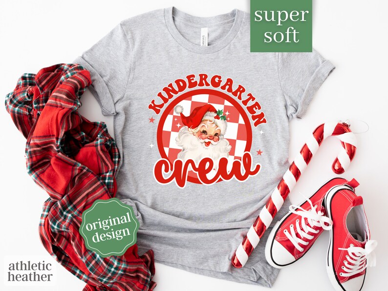 May include: Gray t-shirt with a red and white checkered circle design featuring a cartoon Santa Claus and the text "Kindergarten Crew".