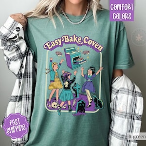May include: A sage green t-shirt featuring a graphic print with the text Easy-Bake Coven. The illustration depicts three figures near a purple oven, a tombstone marked RIP, a skull, and a black cat. The shirt is worn with a black and white plaid shirt.