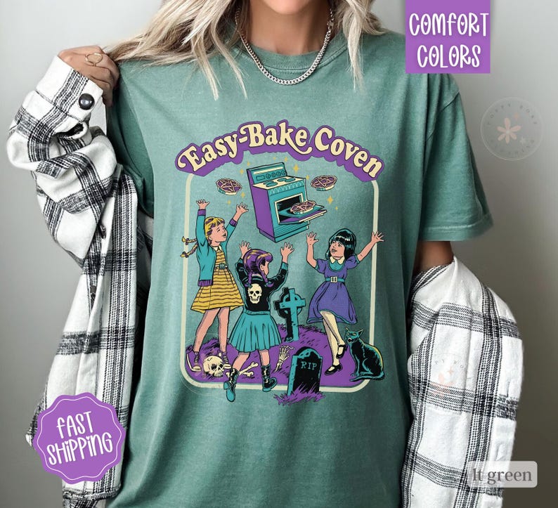 May include: A sage green t-shirt featuring a graphic print with the text Easy-Bake Coven. The illustration depicts three figures near a purple oven, a tombstone marked RIP, a skull, and a black cat. The shirt is worn with a black and white plaid shirt.
