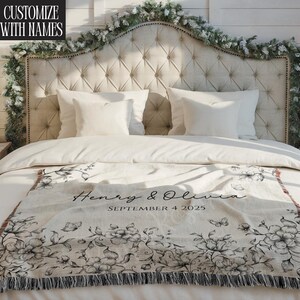 May include: A cream-colored throw blanket with a black floral design and fringe. The blanket is draped over a bed with white bedding and pillows. The text "Henry & Olivia" and the date "SEPTEMBER 4 2025" are printed on the blanket. The text "CUSTOMIZE WITH NAMES" is in the upper left corner.