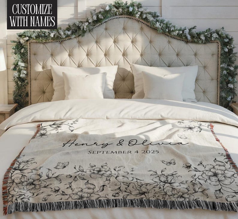 May include: A cream-colored throw blanket with a black floral design and fringe. The blanket is draped over a bed with white bedding and pillows. The text "Henry & Olivia" and the date "SEPTEMBER 4 2025" are printed on the blanket. The text "CUSTOMIZE WITH NAMES" is in the upper left corner.