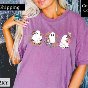 Peut inclure: Un t-shirt violet avec un motif de trois fant&ocirc;mes tenant des fleurs. Les fant&ocirc;mes sont blancs et les fleurs sont roses, orange et jaunes. Le texte "Fast Shipping" est dans le coin sup&eacute;rieur gauche de l'image et le texte "Comfort Colors" est dans le coin sup&eacute;rieur droit de l'image.