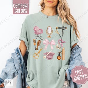 May include: A light green t-shirt with a design of various hair salon tools, including a hair dryer, scissors, combs, a hair dye bowl, a hairspray bottle, and a hair tie. The design is in a watercolor style with pink, gold, and brown accents.