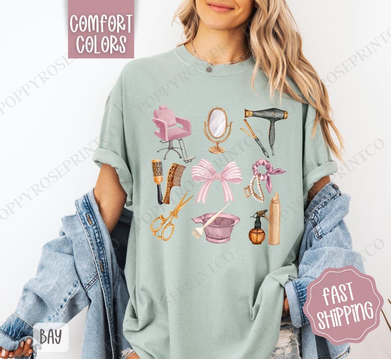 May include: A light green t-shirt with a design of various hair salon tools, including a hair dryer, scissors, combs, a hair dye bowl, a hairspray bottle, and a hair tie. The design is in a watercolor style with pink, gold, and brown accents.
