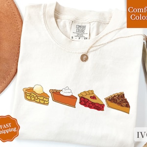 May include: A cream colored t-shirt with a graphic of four different pie slices. The pies are apple, pumpkin, cherry, and pecan. The text "Comfort Colors" is on the tag. The text "IVORY" is on the bottom right corner of the image.
