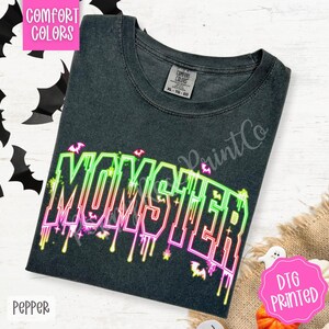 May include: A dark gray t-shirt with the word "MOMSTER" in neon green, pink, and red, with bat graphics. The shirt has a crew neck and short sleeves. The shirt is labeled "Comfort Colors" and "DTG Printed".