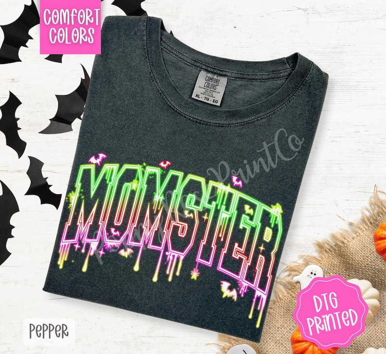 May include: A dark gray t-shirt with the word "MOMSTER" in neon green, pink, and red, with bat graphics. The shirt has a crew neck and short sleeves. The shirt is labeled "Comfort Colors" and "DTG Printed".