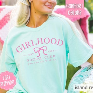 May include: A light blue t-shirt with a pink graphic that says "Girlhood Est 1988 Social Club You Can Sit With Us".