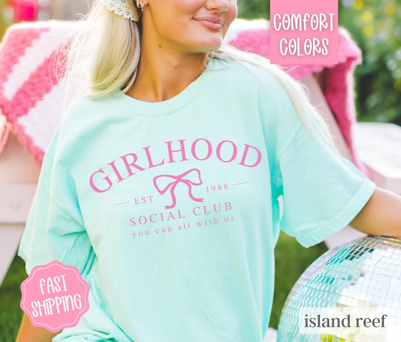 May include: A light blue t-shirt with a pink graphic that says "Girlhood Est 1988 Social Club You Can Sit With Us".