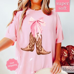 K&ouml;nnte beinhalten: Rosa T-Shirt mit einer rosa Schleife und braunen Cowboystiefeln. Das Shirt tr&auml;gt den Schriftzug "super soft" und "blossom" unten.