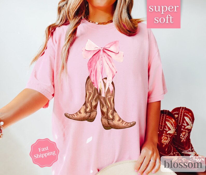 K&ouml;nnte beinhalten: Rosa T-Shirt mit einer rosa Schleife und braunen Cowboystiefeln. Das Shirt tr&auml;gt den Schriftzug "super soft" und "blossom" unten.