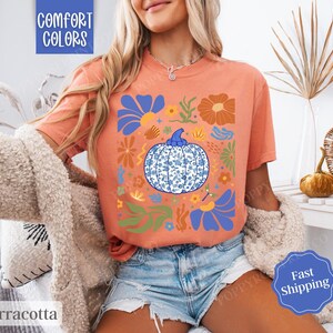 Op de afbeelding: Een terracotta oranje T-shirt met een blauw en wit pompoenontwerp. De pompoen is omringd door bloemen en bladeren in tinten blauw, groen en oranje. De tekst "Comfort Colors" is in wit gedrukt in de linkerbovenhoek van het shirt.
