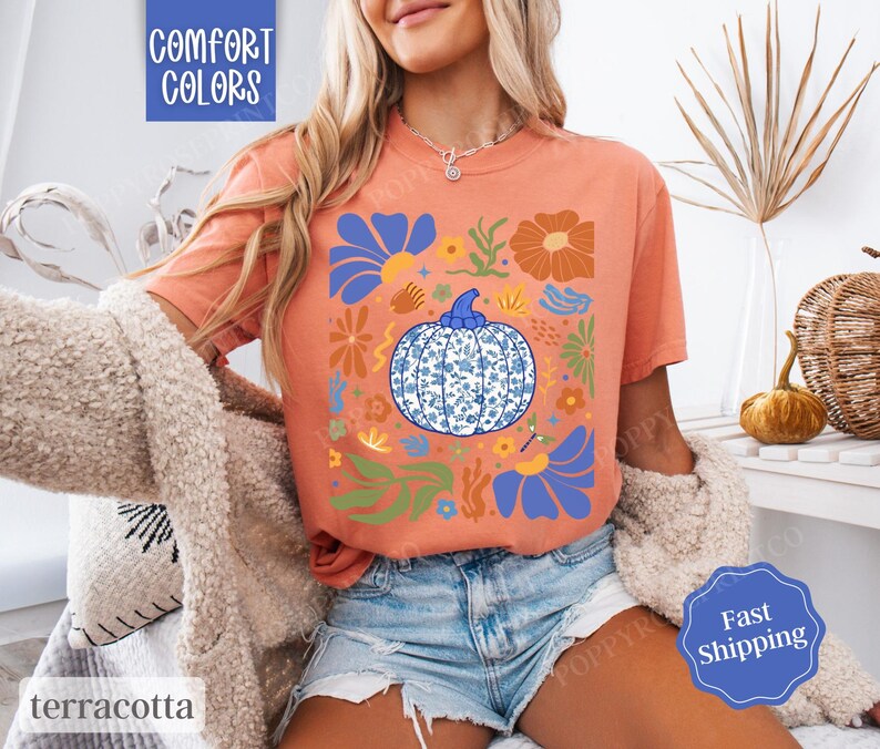 Op de afbeelding: Een terracotta oranje T-shirt met een blauw en wit pompoenontwerp. De pompoen is omringd door bloemen en bladeren in tinten blauw, groen en oranje. De tekst "Comfort Colors" is in wit gedrukt in de linkerbovenhoek van het shirt.