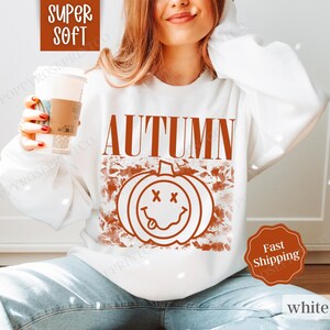 May include: White sweatshirt with a brown graphic of a smiling pumpkin and the word "AUTUMN" in brown text.