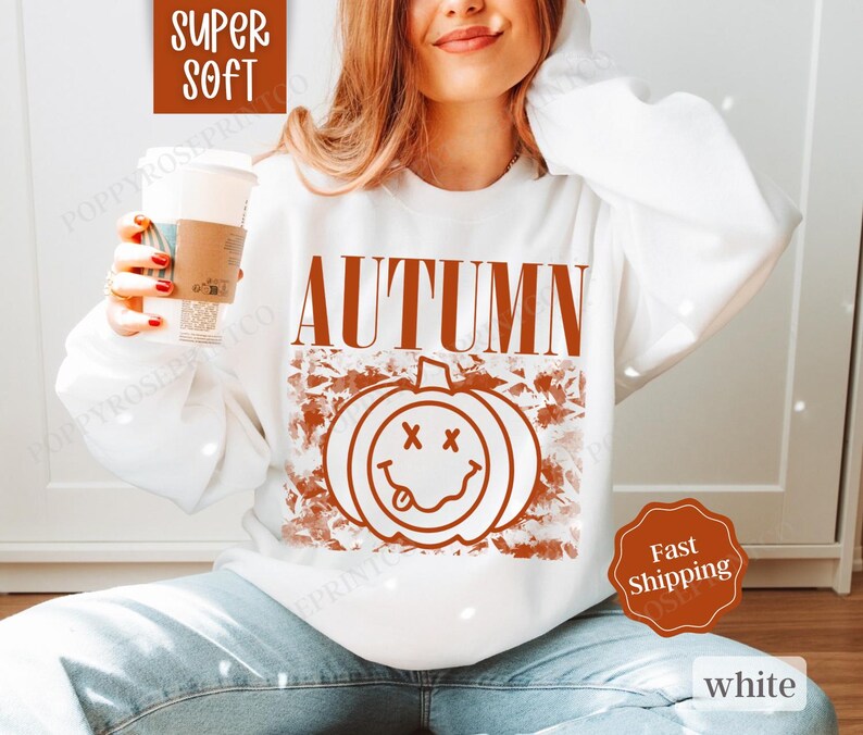 May include: White sweatshirt with a brown graphic of a smiling pumpkin and the word "AUTUMN" in brown text.