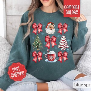 May include: A teal blue sweatshirt with a Christmas theme featuring Santa Claus, a Christmas tree, red bows, and a cup of hot chocolate. The text "Comfort Colors" and "Fast Shipping" are also visible on the sweatshirt.