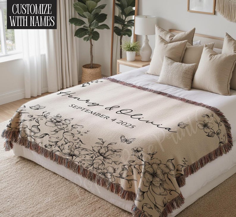 May include: Beige woven throw blanket with a floral design and the names "Henry & Olivia" and the date "SEPTEMBER 4 2025" in black script. The blanket has a fringed edge and is draped over a bed.