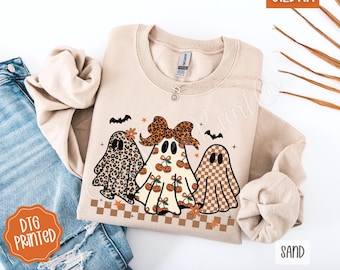 May include: Sand-colored sweatshirt featuring a graphic of three cartoon ghosts with different patterns: leopard print, cherries, and checkered. The design includes bats and flowers. The brand name "GILDAN" is visible in the top right corner. The words "DTG PRINTED" are printed on a sticker.