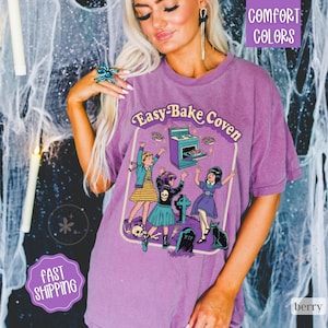 May include: A purple short-sleeve t-shirt featuring a graphic print with the text Easy-Bake Coven. The illustration depicts three figures dancing near a stove, a tombstone, and a black cat. The design includes a skull, bones, and stars.