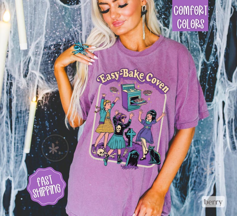 May include: A purple short-sleeve t-shirt featuring a graphic print with the text Easy-Bake Coven. The illustration depicts three figures dancing near a stove, a tombstone, and a black cat. The design includes a skull, bones, and stars.