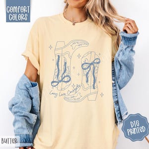 May include: Butter yellow t-shirt with a light blue graphic of two cowboy boots and the text "Long Live Cowgirls." The shirt also has the words "Comfort Colors" and "DTG Printed" printed on it.