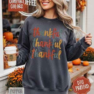 May include: Dark gray sweatshirt with the word "Thankful" embroidered in colorful thread. The word is repeated three times, each in a different color: red, yellow, and teal. The sweatshirt is a comfortable, casual style, perfect for fall.