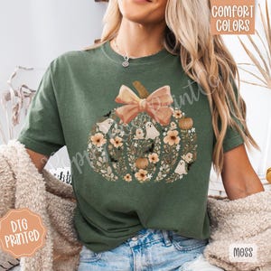 Pu&ograve; includere: T-shirt verde con un design a zucca. La zucca &egrave; decorata con fiori, fantasmi e pipistrelli, e sormontata da un fiocco color pesca. Le parole "Comfort Colors" e "DTG Printed" sono visibili.