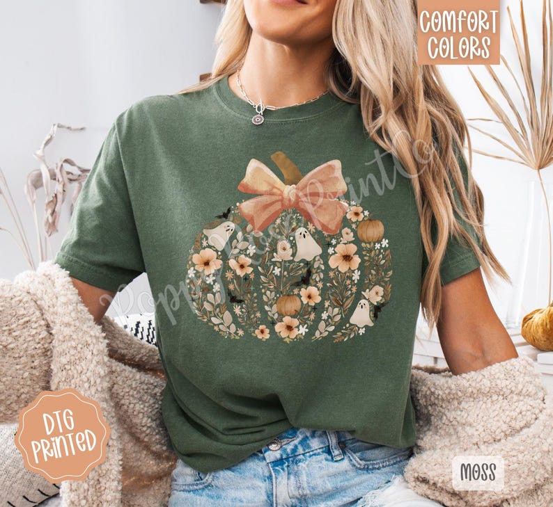 Pu&ograve; includere: T-shirt verde con un design a zucca. La zucca &egrave; decorata con fiori, fantasmi e pipistrelli, e sormontata da un fiocco color pesca. Le parole "Comfort Colors" e "DTG Printed" sono visibili.