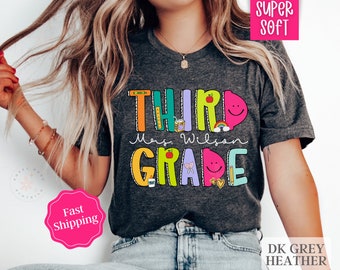 May include: A dark grey heather t-shirt with the text "Third Grade" in colorful letters and a teacher's name. The text is decorated with colorful school supplies and smiley faces.