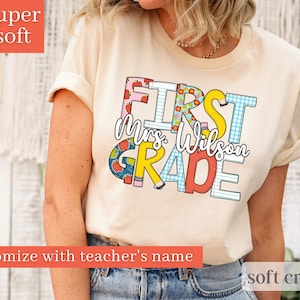 May include: A soft cream coloured t-shirt with the text "FIRST GRADE" in colourful letters with a pattern of apples, pencils, and gingham. The text "Mrs. Wilson" is written in black script below the word "FIRST".