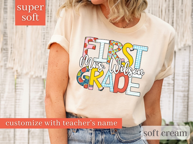 May include: A soft cream coloured t-shirt with the text "FIRST GRADE" in colourful letters with a pattern of apples, pencils, and gingham. The text "Mrs. Wilson" is written in black script below the word "FIRST".