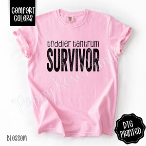 May include: A light pink t-shirt with the words "toddler tantrum SURVIVOR" printed in black. The shirt has rolled-up sleeves and the words "COMFORT COLORS" and "BLOSSOM" are printed on the shirt.