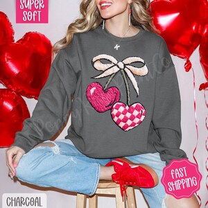May include: Charcoal gray sweatshirt featuring a cherry design with two heart-shaped cherries and a bow. One cherry is solid pink, the other is pink and white checkered. The sweatshirt is paired with blue jeans and red shoes.