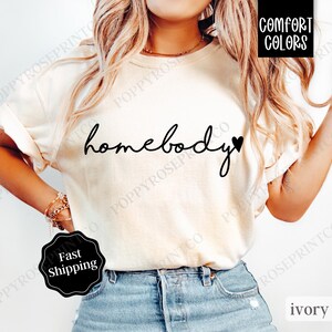 May include: A white t-shirt with the text "homebody" in black script with a small black heart next to it. The shirt is being worn by a person with blonde hair and is paired with blue jeans.