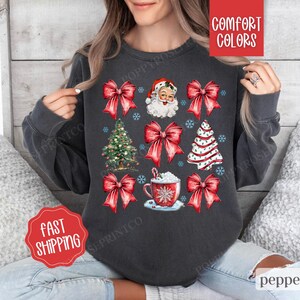 May include: A dark gray sweatshirt with a Christmas theme featuring Santa Claus, Christmas trees, bows, and a cup of hot chocolate. The text "Comfort Colors" is visible in the upper right corner of the image.