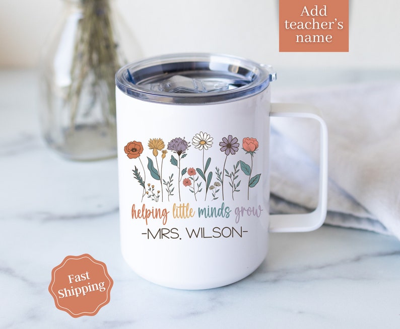Może przedstawiać: Biały kubek podr&oacute;żny z uchwytem i kwiatowym wzorem. Kubek zawiera napis "helping little minds grow" i "-MRS. WILSON-". Projekt zawiera kolorowe kwiaty w kolorze pomarańczowym, ż&oacute;łtym, fioletowym i r&oacute;żowym.