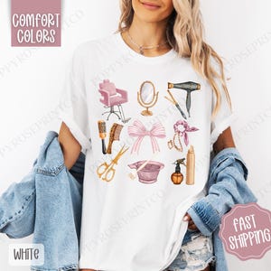 May include: White t-shirt with a pink and gold illustration of hair salon tools, including a hair dryer, scissors, combs, a hair dye bowl, and a hairspray bottle. The text "COMFORT COLORS" is in the upper left corner of the image.