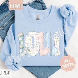 May include: Light blue sweatshirt with the name "Lolli" spelled out in colorful patchwork letters. The letters are made of different fabrics with floral, gingham, and striped patterns.